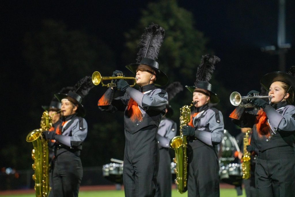 Marching band members in uniforms play brass instruments under stadium lights at night.