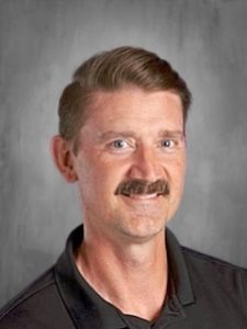 A person with short hair and a mustache, wearing a dark collared shirt, smiles against a gray background.