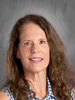 Woman with long brown hair, wearing a blue checkered top, smiling against a gray background.