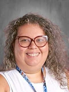 Smiling person with curly hair and glasses, wearing a white top and a lanyard, in front of a gray background.