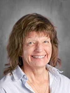 A woman with shoulder-length hair smiles at the camera, wearing a light collared shirt against a neutral background.