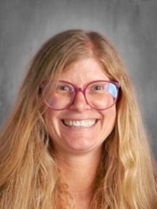 Smiling person with long hair and glasses against a gray background.