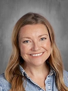 Woman with long light brown hair smiling, wearing a denim shirt, against a gray background.