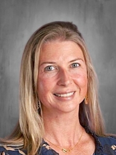 Smiling woman with long blonde hair, wearing a dark patterned blouse and gold earrings, gray background.