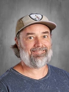 Man with a beard wearing a cap and a blue shirt, smiling against a gray background.