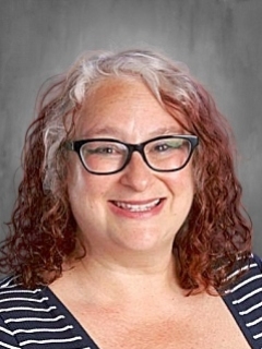 Smiling person with curly hair and glasses, wearing a striped top against a gray background.
