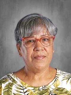 An older woman with short gray hair and glasses, wearing a patterned blouse, against a gray background.
