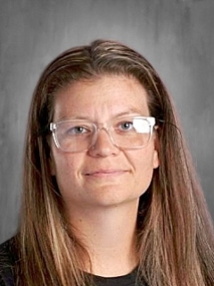 A person with long hair and glasses smiles slightly against a gray background.
