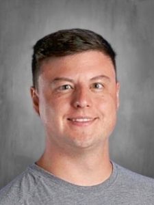 Smiling person with short dark hair wearing a gray shirt against a gray background.