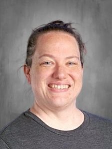 Person with short hair smiling in front of a gray background, wearing a dark shirt.