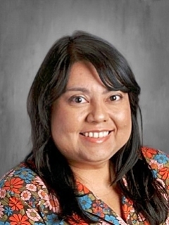 Smiling person with long dark hair, wearing a floral top, against a gray background.