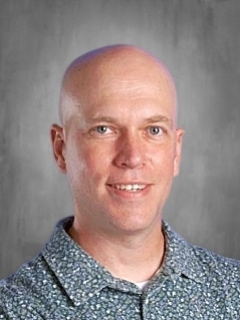 A bald man in a patterned shirt smiles against a gray background.