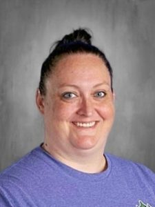 Person with a top bun hairstyle, wearing a purple shirt, smiling at the camera against a gray background.