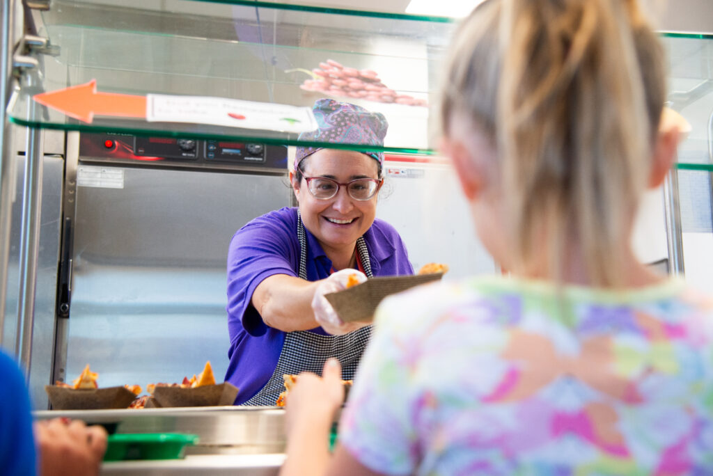 A smiling woman serves food from a cafeteria counter to a child, both engaged in a friendly interaction.