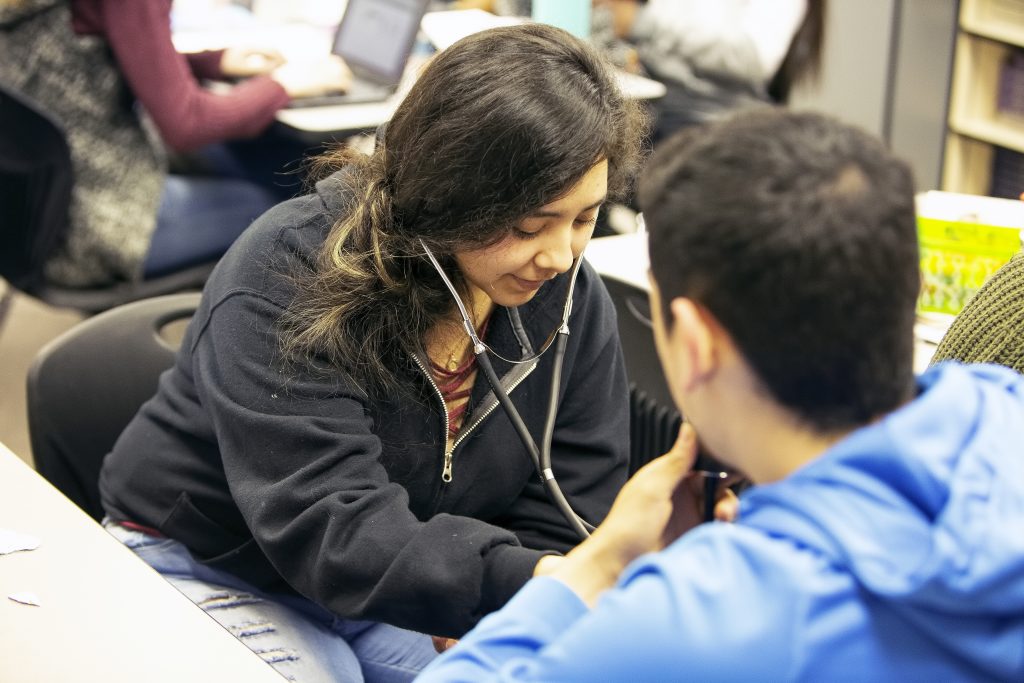 Un estudiante utiliza un estetoscopio mientras conversa con otro estudiante en el aula.