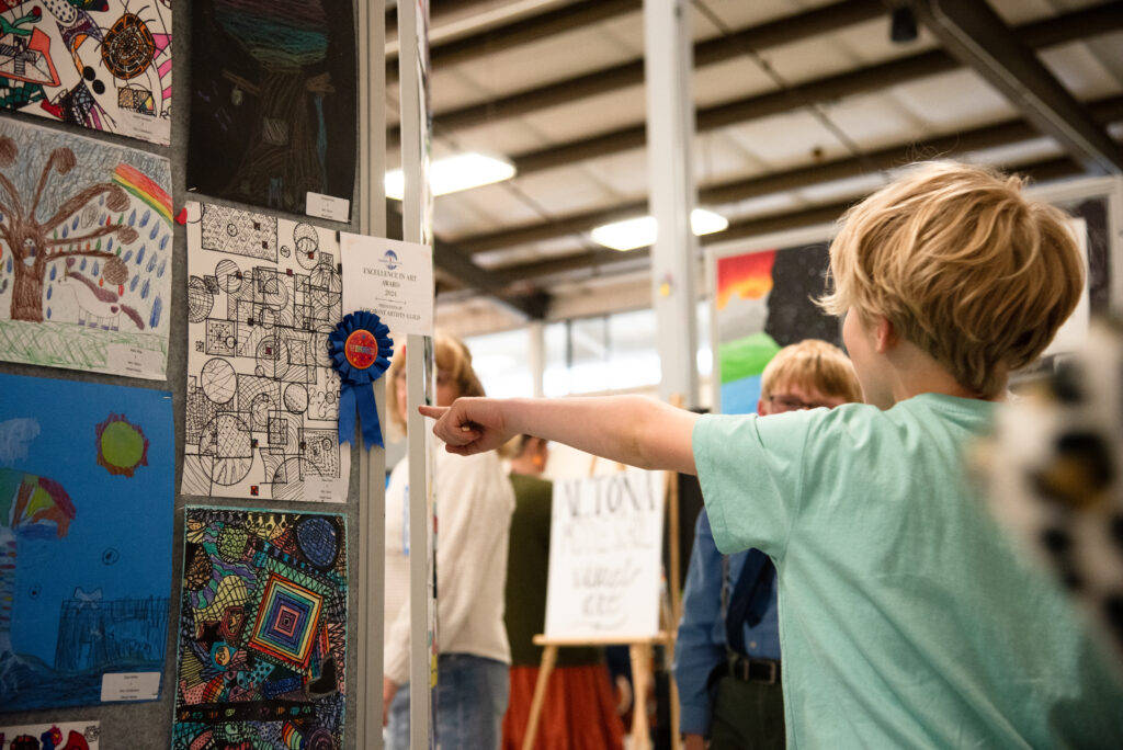 A child points at an award-winning artwork display at an art exhibition, surrounded by other attendees.