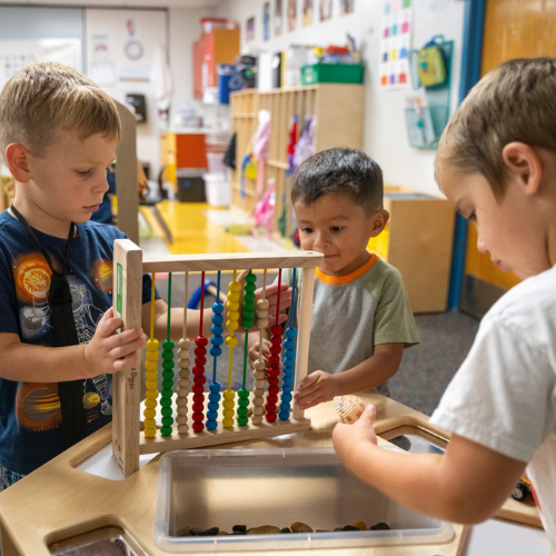 Tres niños pequeños juegan con un ábaco de colores y juguetes para clasificar en un aula luminosa.